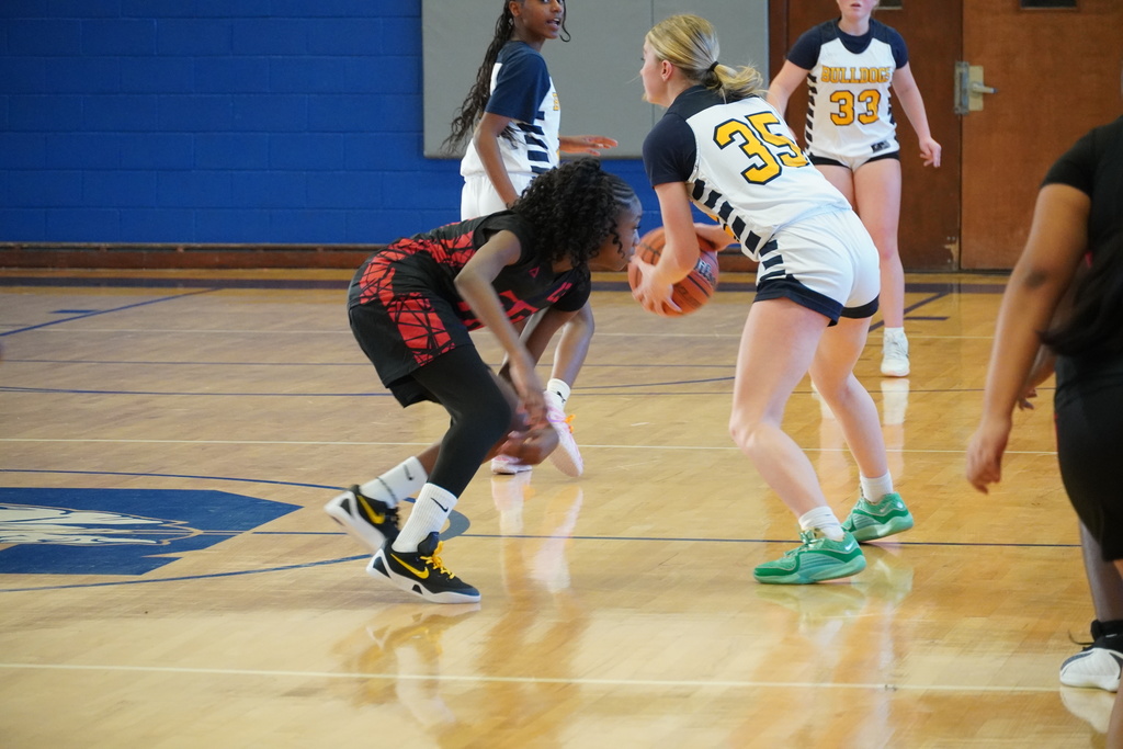 A girls basketball player in a white Bulldogs uniform protects the ball while being closely defended near midcourt during live game action.