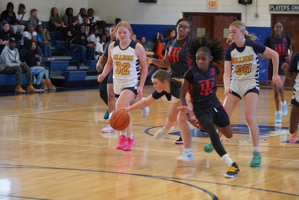 Middle school girls compete for a loose basketball during a game, with players from both teams reaching for the ball as spectators watch from the bleachers.