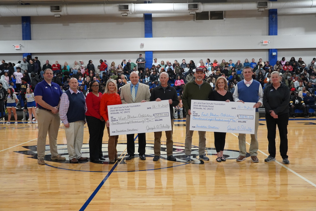 Community members and school staff stand at center court holding ceremonial donation checks during a presentation at a basketball game.