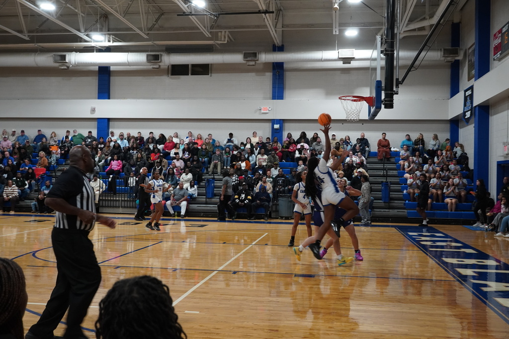 Girls basketball players from both teams jump to contest a shot at the rim as the referee observes from the baseline.