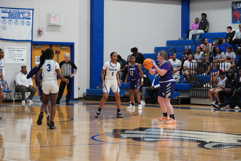 Girls basketball players from the East Bladen Eagles and an opposing team set up on offense and defense near midcourt as a player prepares to pass.