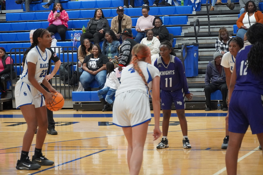 An East Bladen girls basketball player holds the ball on the perimeter while teammates and defenders position themselves during live play, with spectators watching from the bleachers.