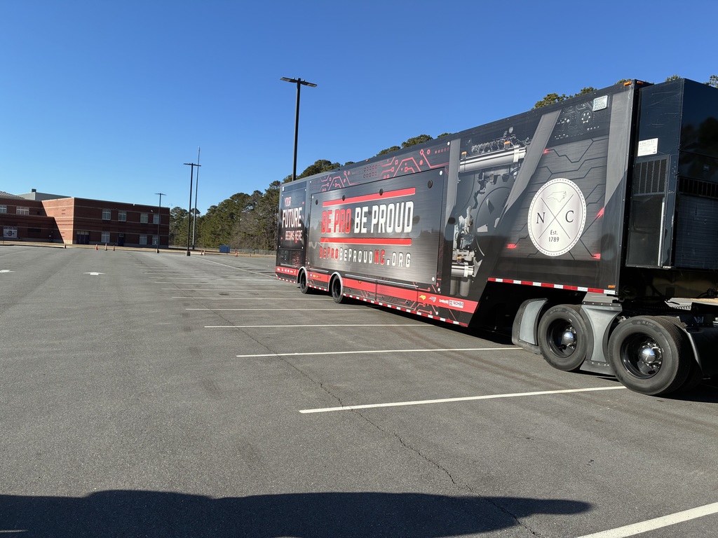 A large black and red semi-trailer truck with “Be Pro Be Proud” graphics is parked in an empty school parking lot under a clear blue sky. The trailer features bold text, circuit-style design elements, and a circular “NC Est. 1789” emblem on the side. A brick school building and tall light poles are visible in the background, with trees lining the edge of the lot.