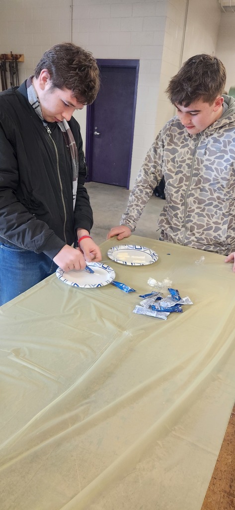 Two students at a table open blue foil wrappers and place the contents onto paper plates during a classroom activity.
