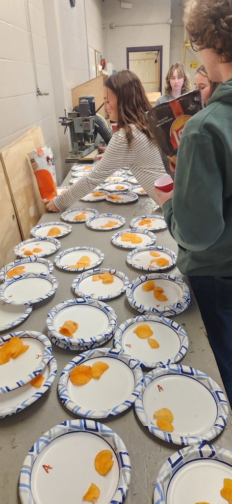 Students prepare snacks by placing potato chips onto rows of paper plates laid out on a long work table.