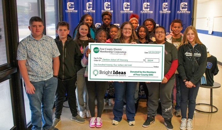 **Alt text:** A group of Clarkton School of Discovery students and staff stand in front of a school banner holding a large Bright Ideas education grant check from Four County EMC for $2,224.
