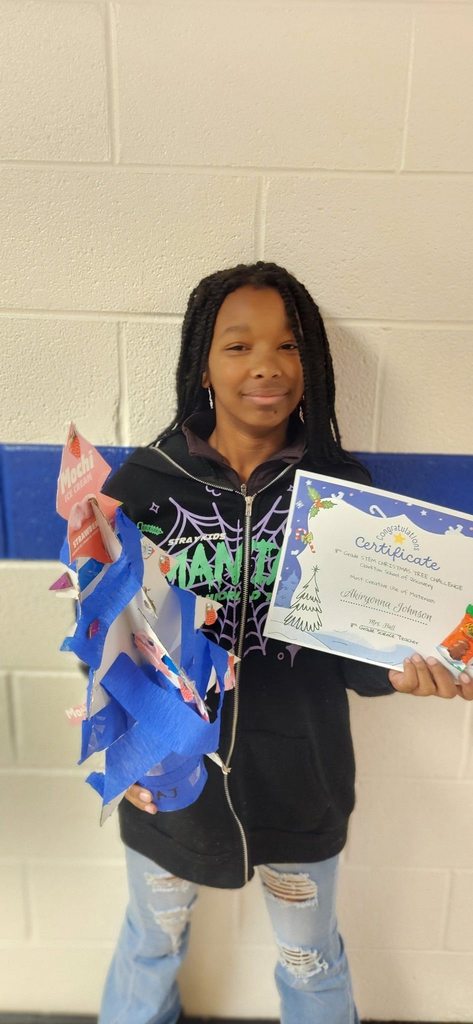 A student holds a recycled Christmas tree made from craft materials and a certificate recognizing creative use of materials.