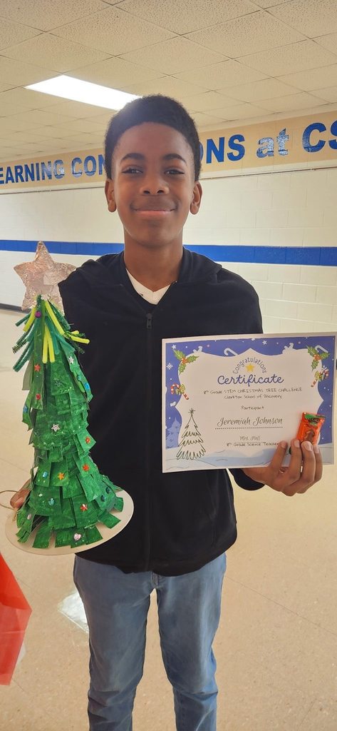 A student stands in a school hallway holding a green recycled Christmas tree and a STEM Challenge participation certificate.