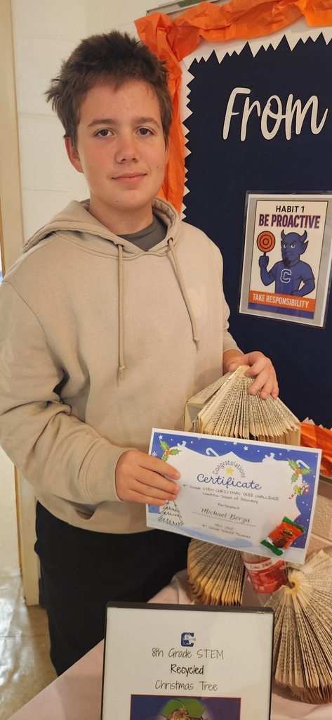 A student stands beside a bulletin board holding a folded-book Christmas tree and a STEM Christmas Tree Challenge certificate.
