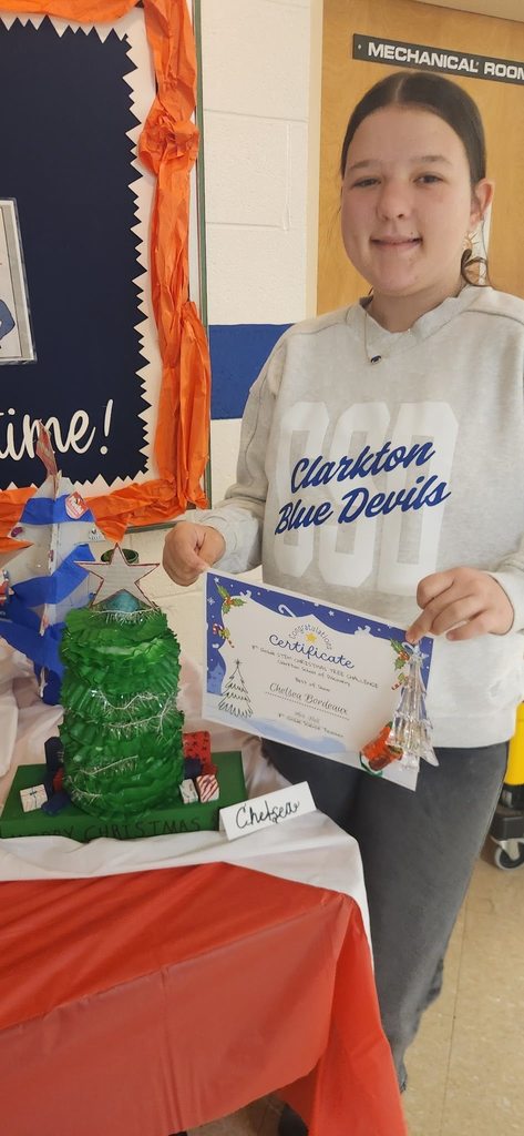A student stands beside a recycled Christmas tree display while holding a certificate from the STEM Christmas Tree Challenge.