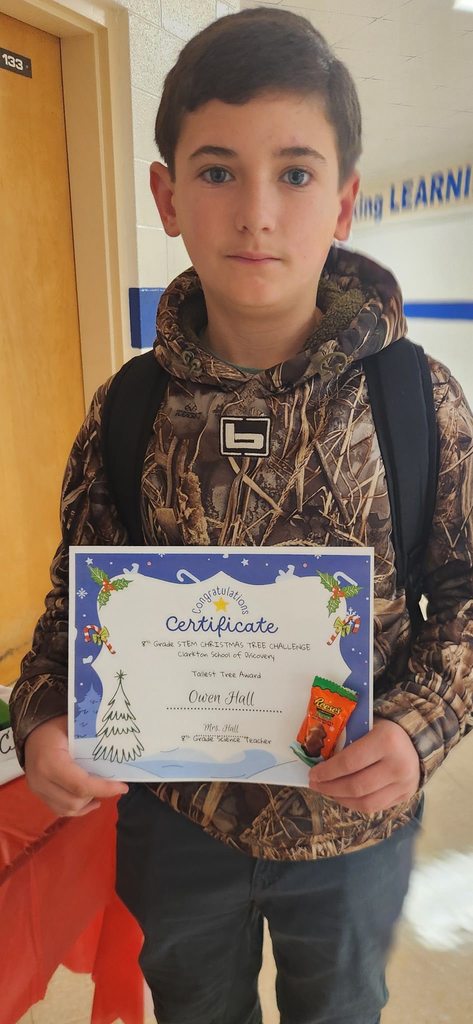A student stands in a school hallway holding a certificate recognizing the Tallest Tree Award from the STEM Christmas Tree Challenge.