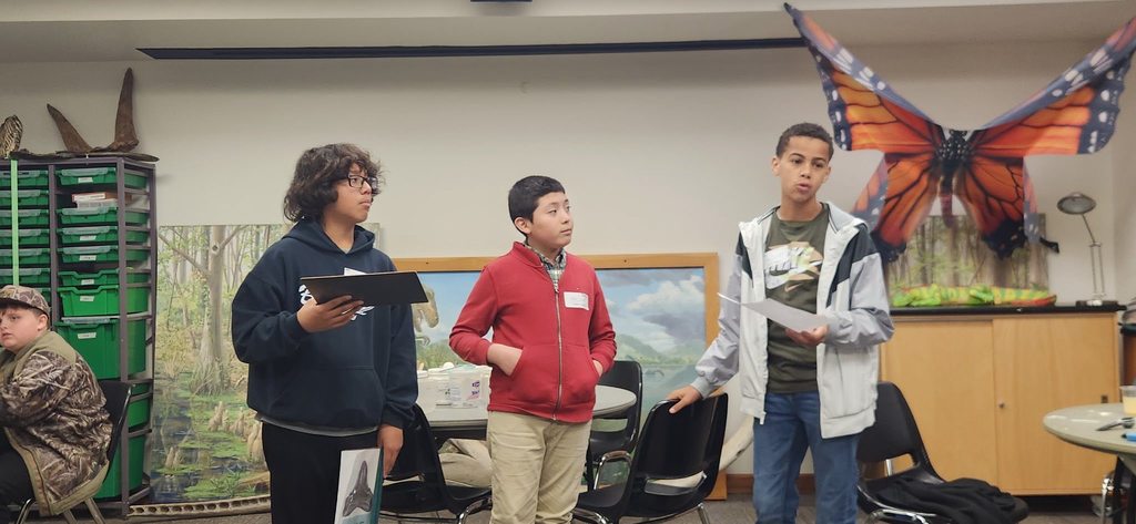 Three students stand in a classroom presenting and discussing a science activity while holding papers.