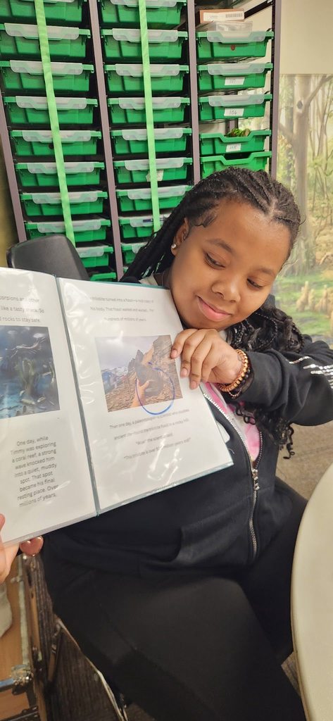 A student sits indoors holding open a science book and pointing to an image while reading.