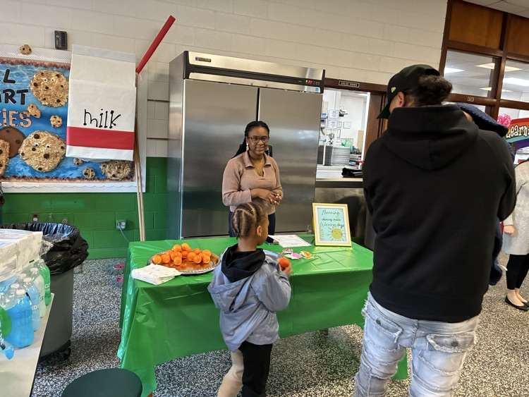 young student and adult stand in front of table with green tablecloth talking to school staff member