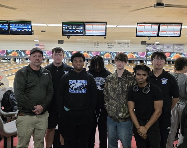 one male coach and six male bowlers standing in the bowling alley