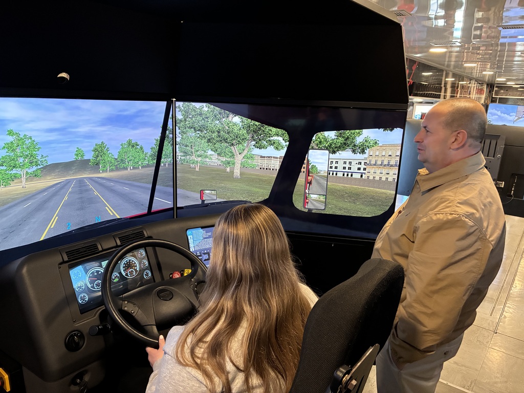 The scene is inside a full-size or semi-enclosed truck cab simulator, complete with a steering wheel, dashboard gauges, side mirrors, and driver’s seat. A person is seated in the driver’s seat, hands on the wheel, actively “driving.” Another adult is standing just behind and to the side, watching closely—probably instructing, supervising, or observing performance. The windshield and side windows are actually large screens showing a virtual roadway with lanes, trees, and buildings, simulating real driving conditions. The environment looks like an indoor training facility or exhibit space (polished floor, overhead lighting, reflective surfaces).
