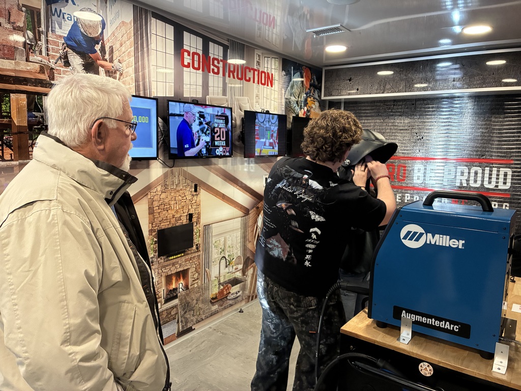 A welding simulation station (the blue Miller AugmentedArc unit) set up on a workbench. A young person wearing a welding helmet, actively practicing or being guided through a weld. Another person close by (likely an instructor or mentor) helping position the helmet or giving instruction. An older man observing from the side, watching attentively. The space itself looks like a construction or trades training trailer / classroom, with big wall graphics, screens showing instructional content, and the word “CONSTRUCTION” prominently displayed. Overall vibe: education + workforce training, probably introducing welding or skilled trades in a safe, simulated environment before real welding.