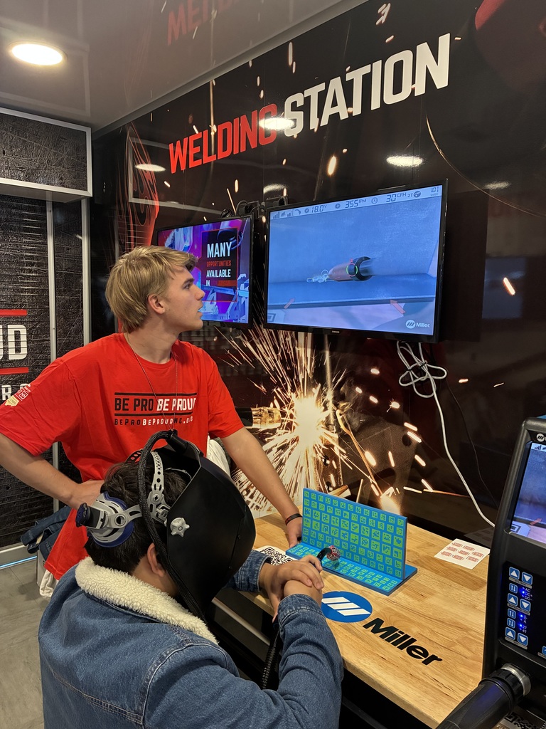 A branded “Welding Station” booth (Miller logo on the table). One person wearing a welding helmet, holding a simulated torch and practicing on a small mock workpiece. Another person in a red shirt standing beside them, watching the real-time simulation on a wall-mounted screen. The screen shows a virtual weld bead, with stats like angle, speed, and position—typical training feedback. The whole setup feels like a career fair, trade expo, or skills training event, focused on teaching welding safely without real sparks.