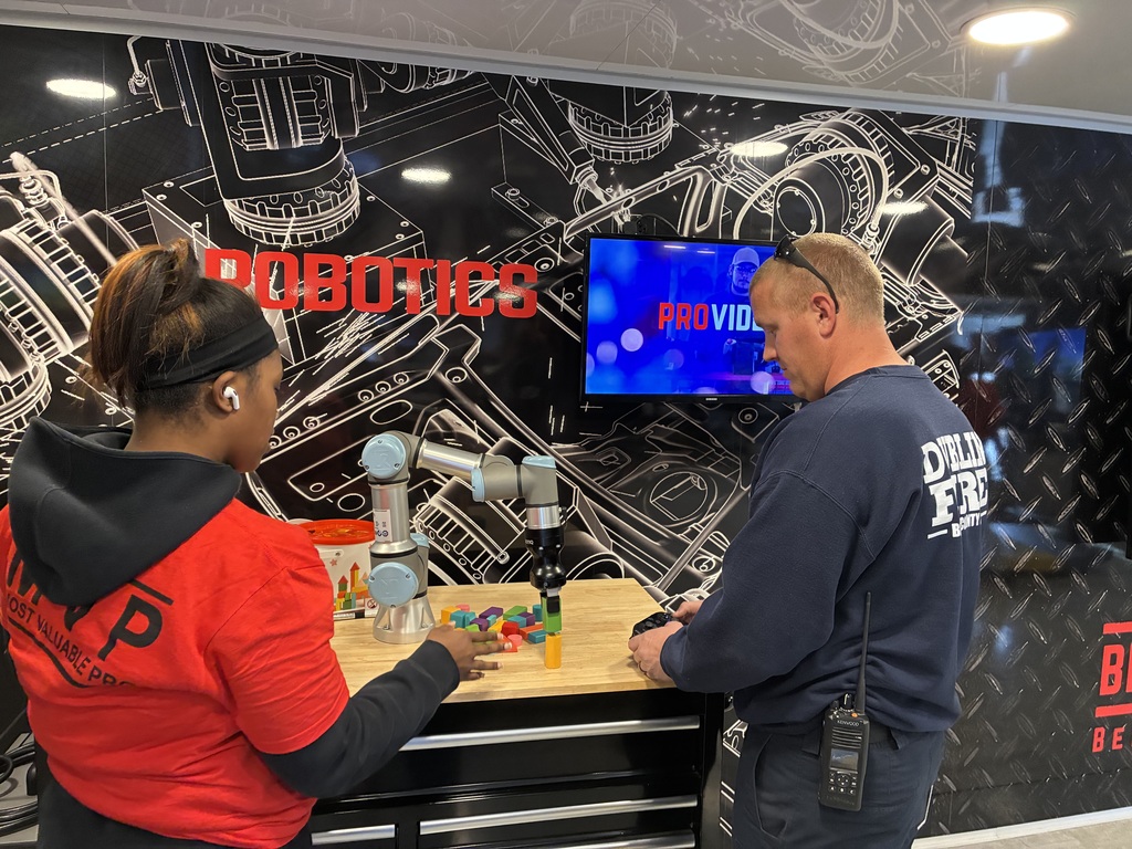 two people standing at a workbench with a collaborative robot arm (a cobot) in front of them. The robot appears to be stacking or manipulating colorful wooden blocks on a small table. One person (left) looks like they’re guiding or demonstrating the task, while the other (right) is watching closely, maybe learning or supervising. Behind them is a big wall graphic with mechanical schematics and the word “ROBOTICS” in bold red letters, plus a screen playing a promo or demo video—definitely a tech showcase vibe, possibly at a training center, event, or mobile robotics lab.