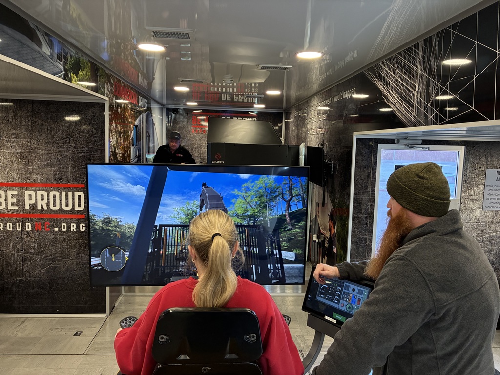 Inside a mobile training trailer, a person sits in a simulator chair facing a large screen that displays a forestry logging simulation with heavy machinery and trees. An instructor stands to the right, operating a touchscreen control panel, while another person observes from the back. The interior walls feature industrial graphics and safety-themed signage.
