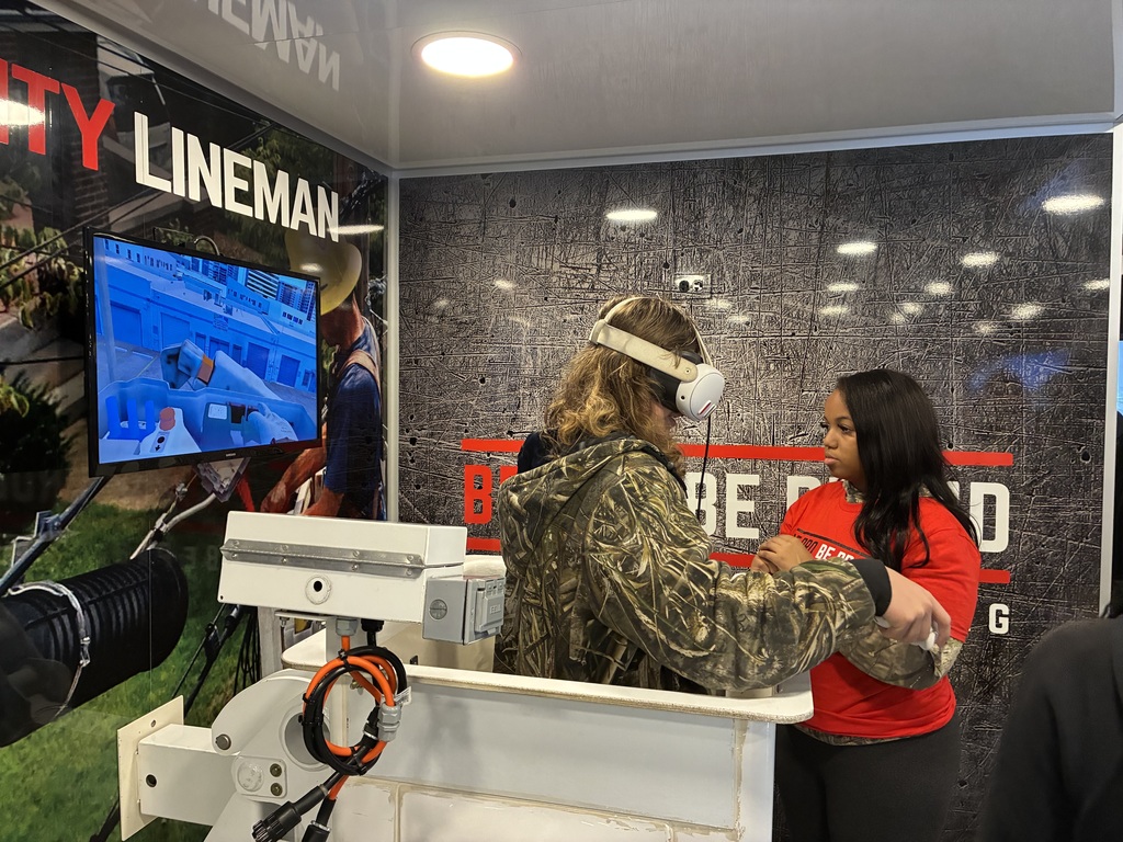 A participant is wearing a VR headset and standing at a bucket-truck simulator, practicing tasks you’d do as a power line worker. A trainer/instructor stands in front of them, guiding or coaching them through the exercise. On the left, a monitor shows the VR view—it looks like a virtual worksite with tools and equipment, so the trainee can see exactly what they’d experience in the field. The booth branding includes “Lineman” and visuals of real utility work, suggesting this is designed for job training, safety practice, or workforce development. The setup mimics real conditions but in a controlled, safe environment, which is great for learning without real-world risk.