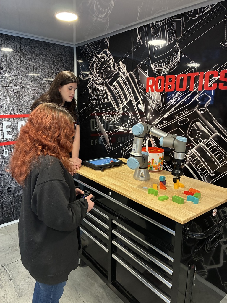 Two people are standing at a workbench with a small collaborative robotic arm (the kind used for teaching and light industrial tasks). The robot is set up to pick and place colorful wooden blocks that are spread out on the tabletop. One person is holding a game-style controller, likely driving or programming the robot, while the other is watching closely and assisting. Behind them, the wall is covered with a high-contrast, blueprint-style mural of mechanical components, with the word “ROBOTICS” in bold red letters—very workshop-meets-tech-showroom vibes. There’s also a tablet on the bench that appears to be running the robot’s interface, plus a container holding additional blocks or parts. Overall, it feels like an interactive learning station—something you’d see at a STEM lab, robotics class, or tech expo—focused on teaching robot control, coordination, and automation in a very approachable, hands-on way