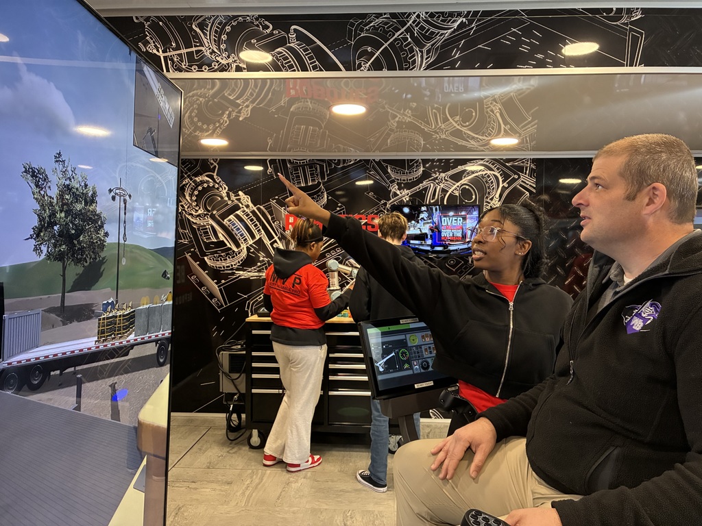In the foreground, a woman in a black hoodie is pointing up at a large vertical screen on the left that shows what looks like a crane or heavy-equipment simulation (a tree, load, and trailer are visible). A man sitting beside her is watching closely, holding a controller, like he’s the one actually “driving” in the simulator. Behind them, a couple of other people in red shirts are working at a tool bench and another screen, which gives the whole place a training or trade-show vibe. The walls are decorated with technical, blueprint-style graphics of machinery, reinforcing the industrial / equipment theme. Overall, it feels like someone is explaining how to operate the crane or pointing out what to do next in the simulation—very coach-and-trainee energy.