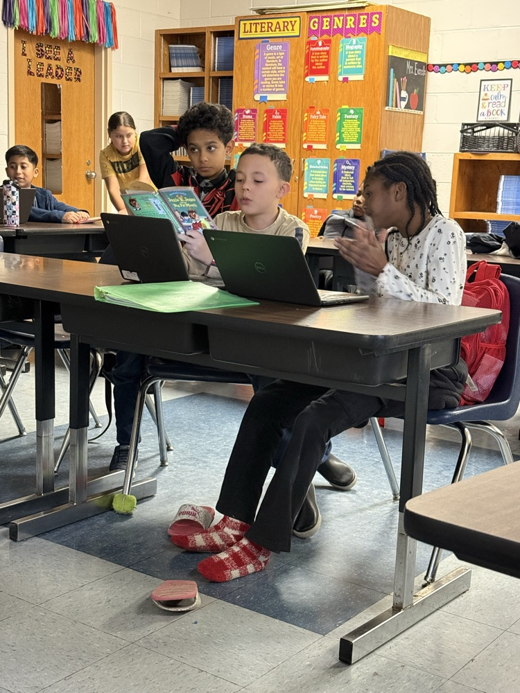 Students sit together at desks reading a book aloud while using laptops, collaborating and listening to one another in a classroom with literacy posters on the walls.