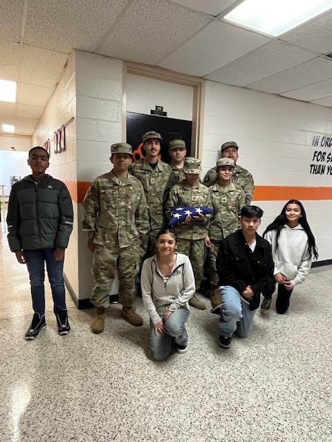 A group of East Bladen High School JROTC cadets in military uniforms stand in a school hallway with Elizabethtown Middle School students. One cadet holds a neatly folded American flag, symbolizing respect and proper flag etiquette. Students pose proudly alongside the cadets during the flag-handling training.