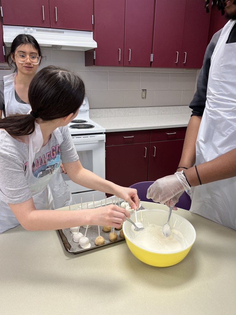 A student wearing glasses and an apron watches closely as another student stirs a bowl in front of a kitchen stove and oven
