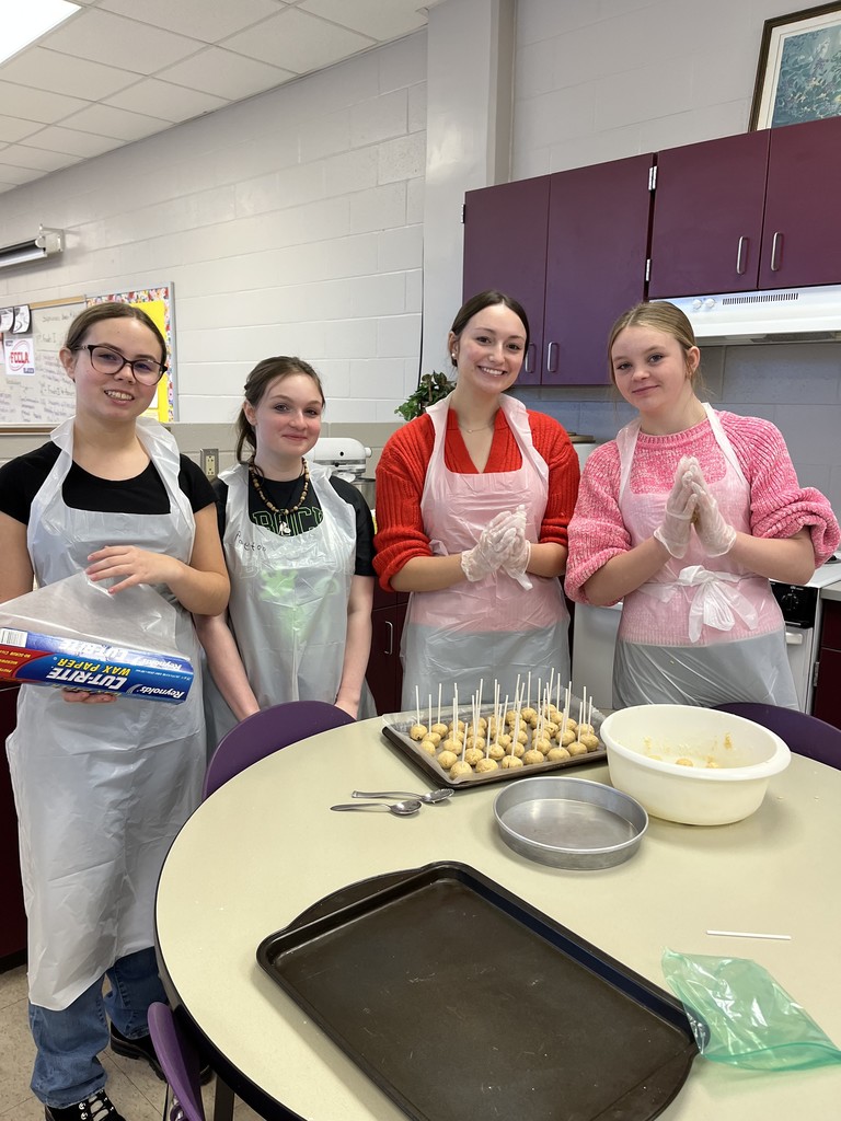 Four students in a classroom kitchen setting standing around a circular table, smiling and posing together during a food preparation activity