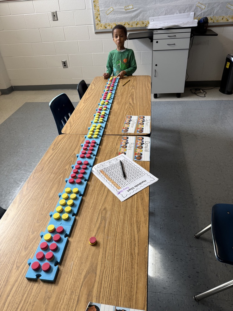 A young student stands at a classroom table using colorful math manipulatives arranged in a long line to practice counting from 1 to 100 and counting by tens. Number charts and worksheets are on the table as he works independently.