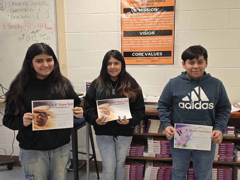 Three Elizabethtown Middle School students pose in a classroom holding Honor Roll and Perfect Attendance certificates beneath the school’s mission and core values poster.