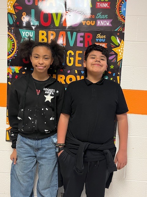 Two Elizabethtown Middle School students stand side by side in a school hallway in front of a colorful inspirational poster. Both students are smiling at the camera, wearing black tops, and representing ETMS Students of the Month.