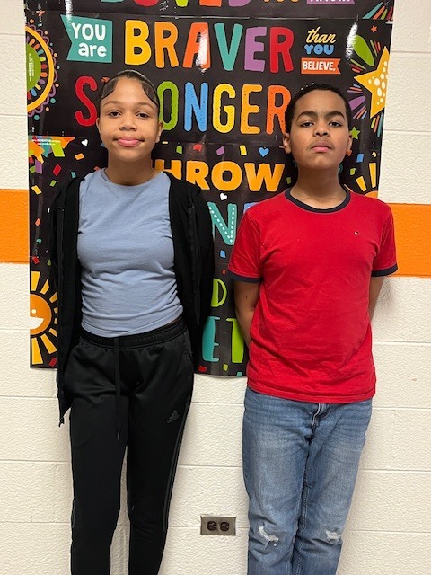 Two Elizabethtown Middle School students stand side by side in a school hallway in front of a colorful inspirational poster. One student wears a blue shirt and black jacket, and the other wears a red shirt and jeans, both facing the camera and representing ETMS Students of the Month.