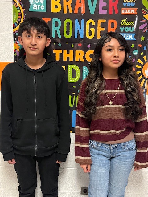 Two Elizabethtown Middle School students stand side by side in a school hallway in front of a colorful inspirational poster. One student wears a black hoodie and pants, and the other wears a striped sweater and jeans, both facing the camera and representing ETMS Students of the Month.