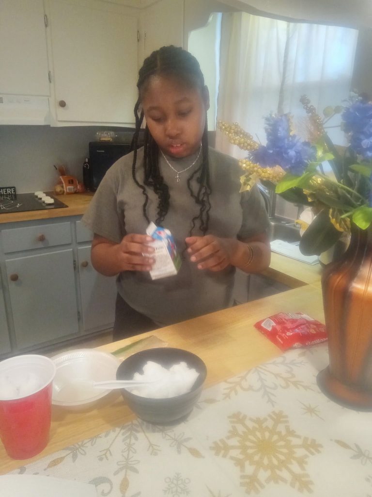 A student in a grey t-shirt stands in a kitchen, holding a small milk carton over a bowl. Kitchen items, flowers, and a red bag of ingredients are visible on the counter.