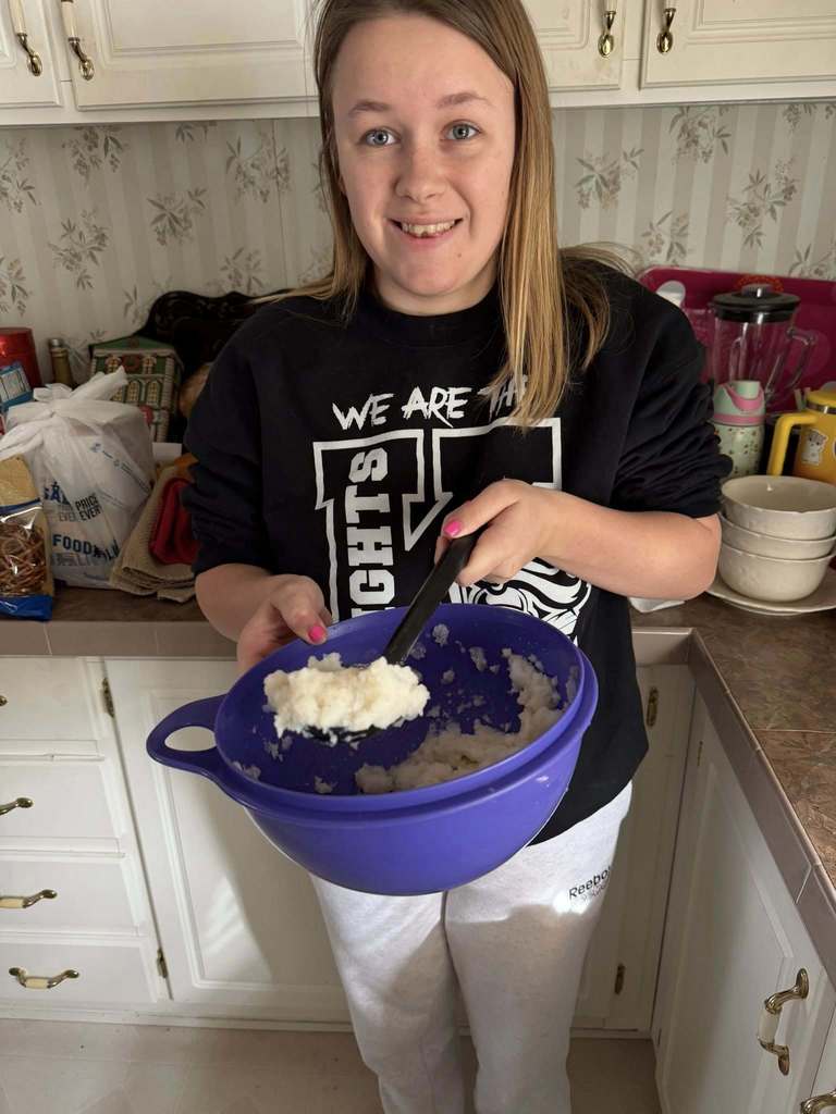 A student in a "Knights" sweatshirt uses a large spoon to mix snow cream in a purple bowl on a kitchen counter.