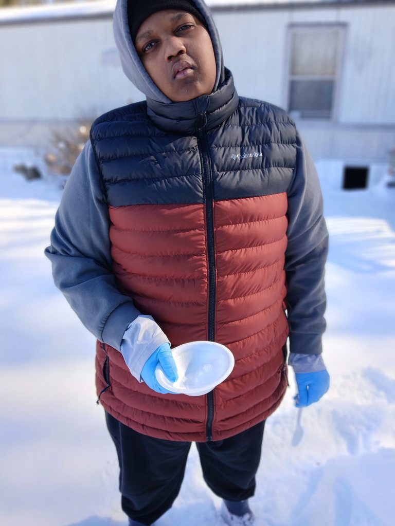 A student in a blue and orange puffer vest and blue gloves holds a white bowl of snow while standing outside in a snowy landscape.