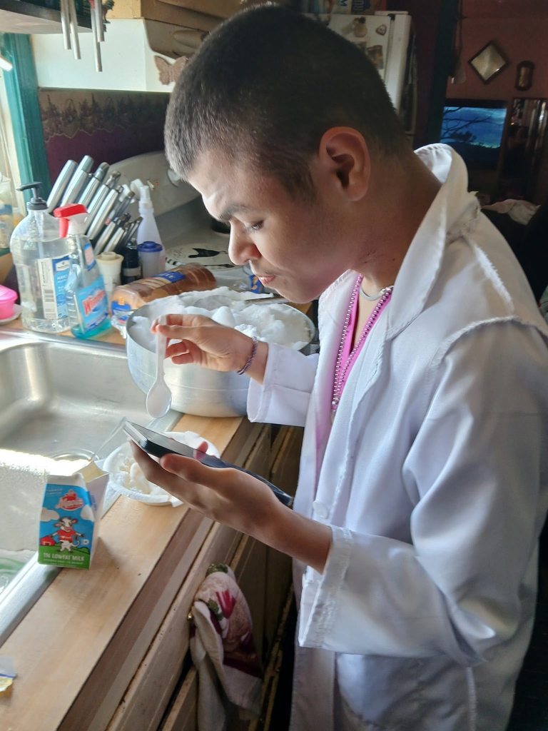 A student in a white lab coat looks down at a phone while preparing snow cream in a kitchen. They hold a white spoon over a bowl of snow, with milk and containers on the counter.