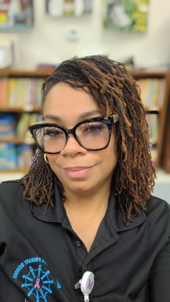 Portrait of Mrs. Pridgen, the school guidance counselor, smiling in front of bookshelves in the school library. She is wearing glasses and a black school shirt, representing National School Counseling Week and celebrating the role of school counselors in supporting student success.