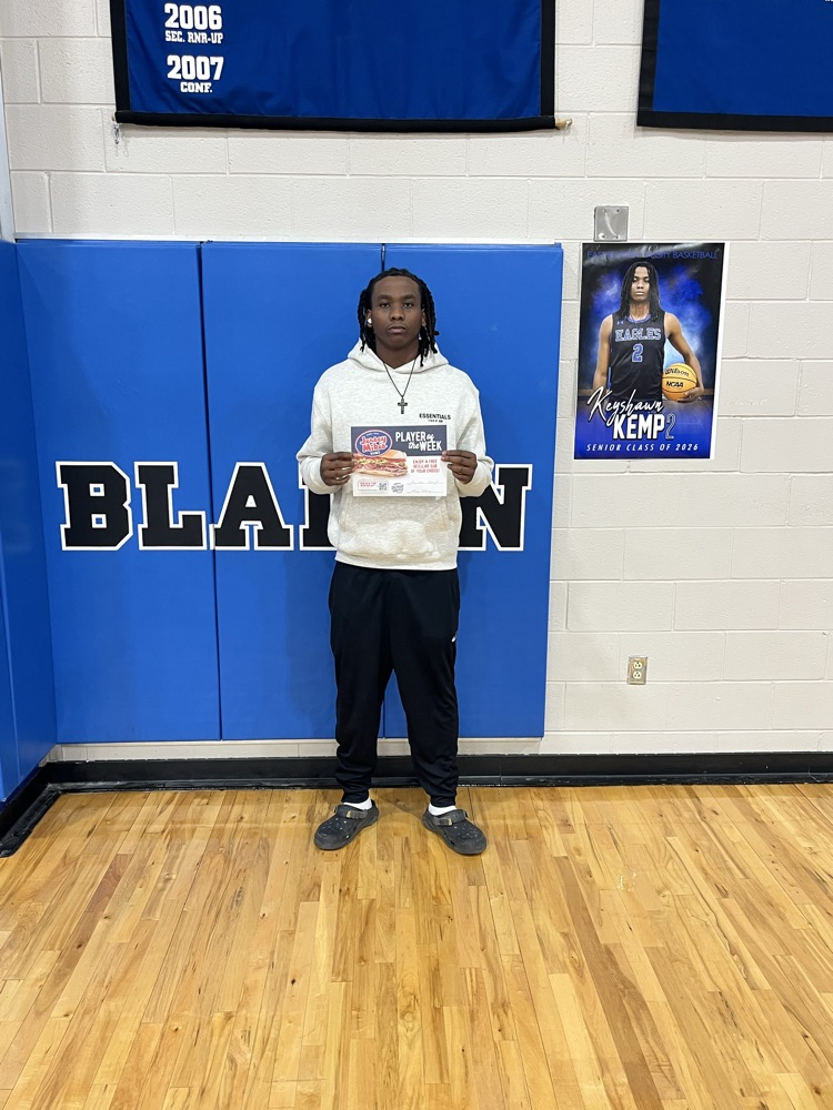 Key Kemp stands in a school gym in front of blue wall padding, holding a “Player of the Week” certificate.