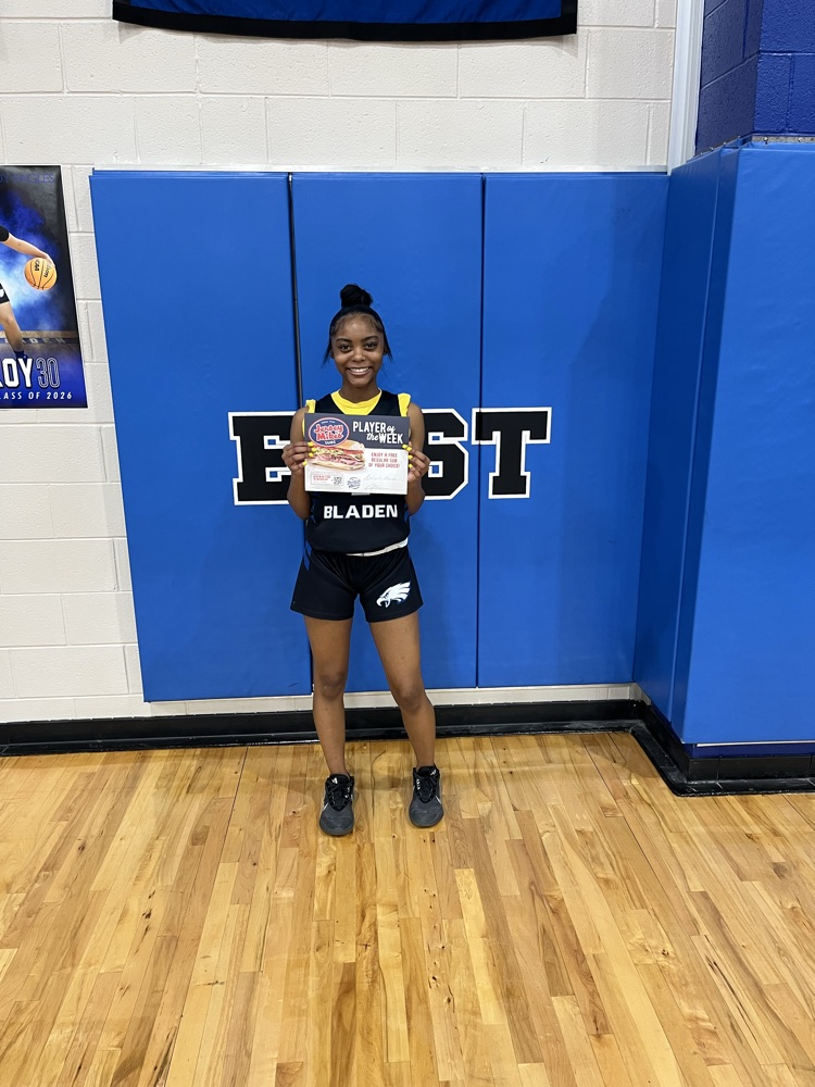 Aaliyah stands in a school gym wearing an East Bladen uniform, smiling and holding a “Player of the Week” certificate.