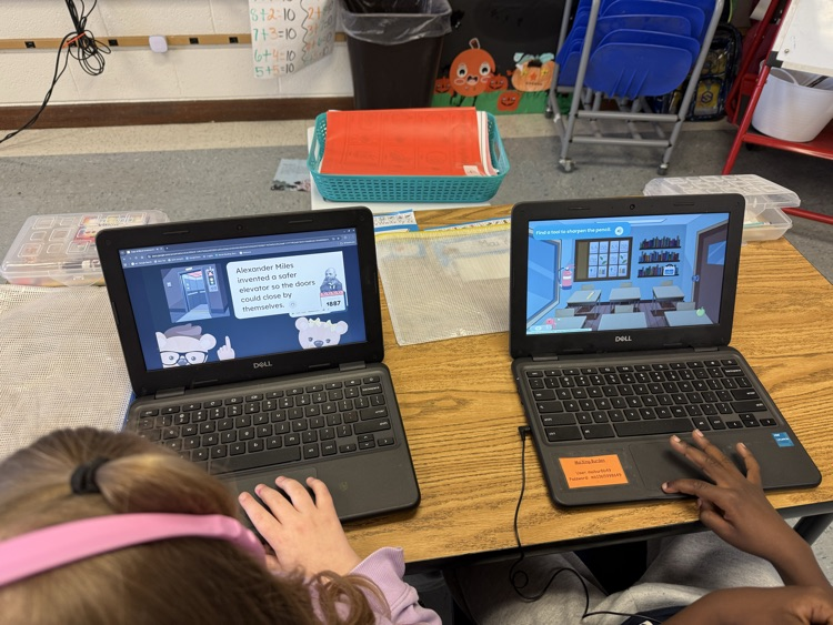 First grade students sit at classroom tables using laptops and headphones while completing an online scavenger hunt about African American inventors. Digital clues and interactive activities are displayed on their screens as they research and learn.
