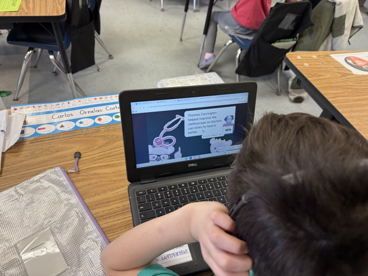 First grade students sit at classroom tables using laptops and headphones while completing an online scavenger hunt about African American inventors. Digital clues and interactive activities are displayed on their screens as they research and learn.