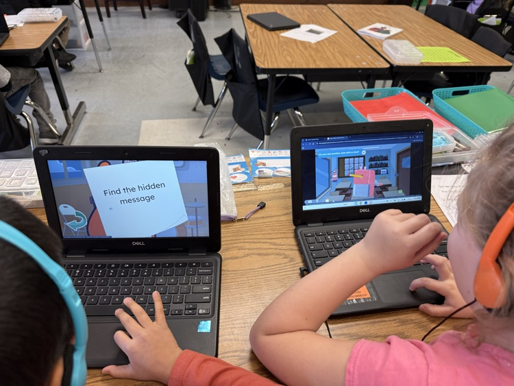 First grade students sit at classroom tables using laptops and headphones while completing an online scavenger hunt about African American inventors. Digital clues and interactive activities are displayed on their screens as they research and learn.