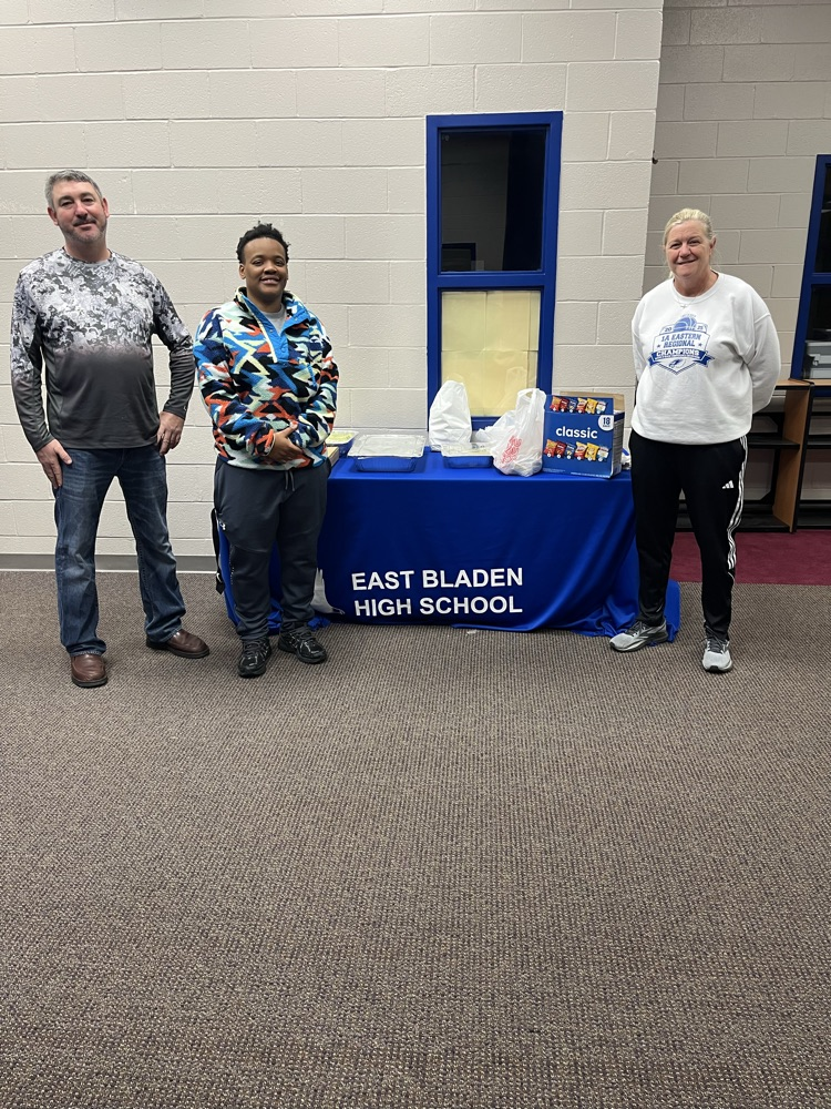a man, and two women stand beside a table with a blue tablecloth reading East Bladen high school and on it is food