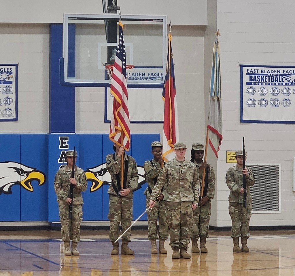 6 students in JROTC uniforms are presenting colors (flags) in a gym. There is an Eagle on the wall in the background.