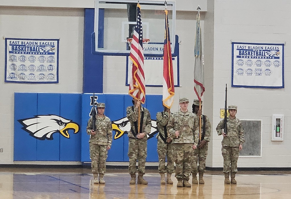 6 students in JROTC uniforms are presenting colors (flags) in a gym. There is an Eagle on the wall in the background.