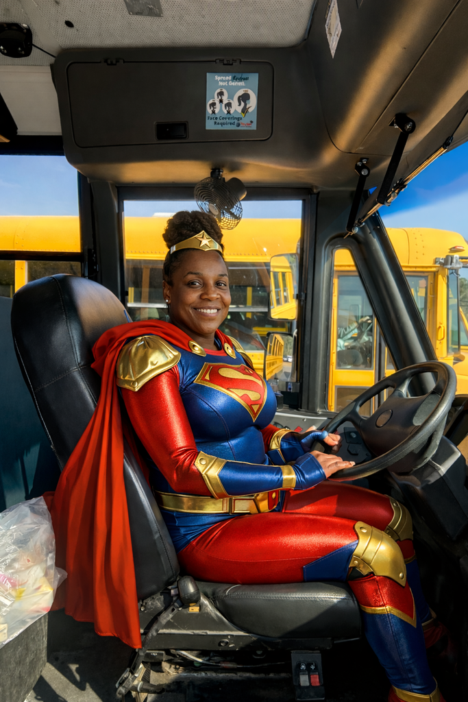 A smiling woman dressed in a red, blue, and gold superhero costume with a red cape sits in the driver’s seat of a school bus, holding the steering wheel. Yellow school buses are visible through the windows behind her, and sunlight fills the bus interior.